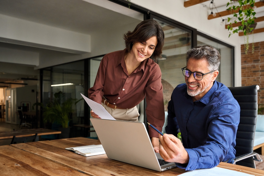 alt Two colleagues smiling and collaborating over a laptop at the office